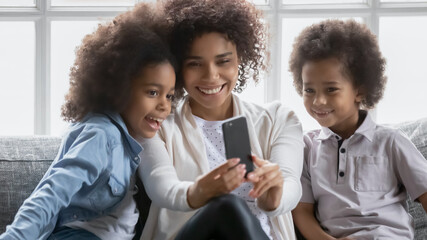 Close up overjoyed African American woman with kids taking selfie, holding smartphone, looking at screen, smiling mother with adorable son and daughter using phone, video call, enjoying leisure time