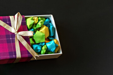 Colored gift box with satin bow with origami paper stars on black background. Gifts for the holidays.