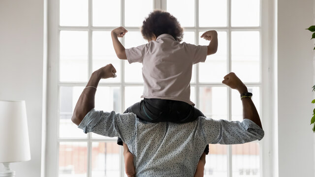 Rear view African American father and little son sitting on dad shoulders showing biceps, strength, healthy lifestyle and fit concept, playing active game, two superheroes, support and care