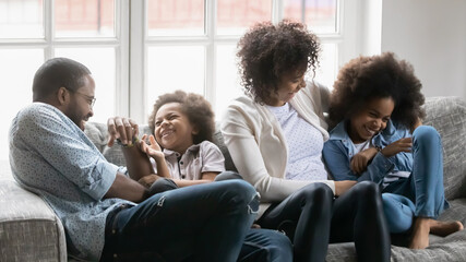 Close up overjoyed African American parents with kids tickling, having fun, sitting on cozy couch at home, happy mother and father with little son and daughter cuddling, involved in funny activity