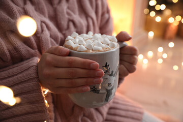 Woman holding cup of hot drink with marshmallows indoors, closeup. Magic Christmas atmosphere