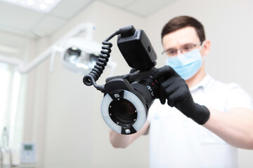 The dentist takes a picture on the camera during the appointment. Doctor in protective mask out of focus. A camera with a ring flash in the hands of a doctor. Doctor's hands in protective black gloves