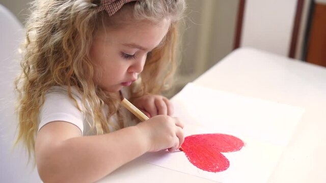 Little blonde girl sits at the table, draws a red Valentine's heart. DIY valentine's day card.