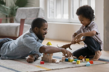 Close up happy African American father and son playing with toys together, sitting on warm floor with underfloor heating, constructing with wooden blocks, family enjoying leisure time at home