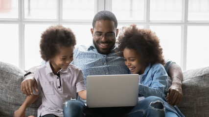 Close up smiling African American father with kids using laptop, looking at screen, sitting on couch at home, happy dad with little daughter and son watching video, shopping or playing game
