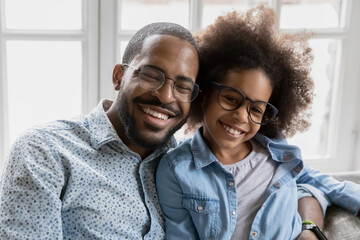 Head shot portrait happy African American father and adorable daughter wearing glasses hugging,...