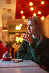 Woman in sweater sitting in restaurant with cup of fruit tea and looking out the window