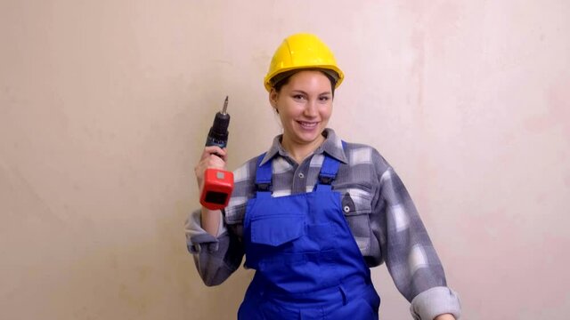 A young beautiful girl construction worker in a yellow helmet and blue uniform dances with a screwdriver in her hands against a gray wall. Girl repairman
