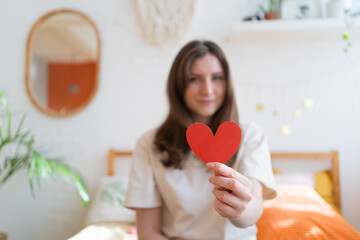 Give love on Valentine's Day. Red paper heart in the hand of a young woman in a warm room