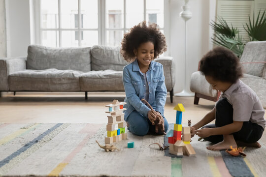 Happy African American boy and girl playing with toy dinosaurs and wooden blocks cubes in living room, sitting on warm floor with underfloor heating, little siblings having fun at home together