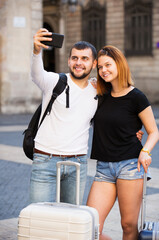 Young couple of tourists with luggage taking photo on mobile phone at street