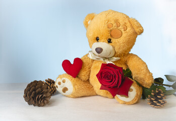 A funny character Teddy Bear holds a red heart and a scarlet rose in his paws on a light background with bokeh effect. The concept of Valentine's Day and love.