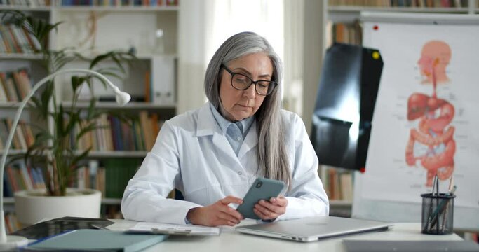 Mature female doctor in glasses touching smartphone sreen. Good looking woman in white professional coat using phone while sitting at table in medical office. Concept of internet