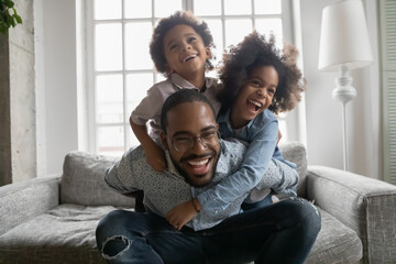 Close up overjoyed African American man wearing glasses piggy backing kids, sitting on couch at...