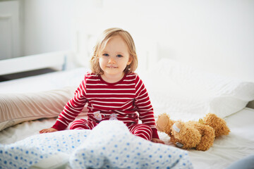 Happy toddler girl in striped red and white pajamas sitting on bed right after awaking