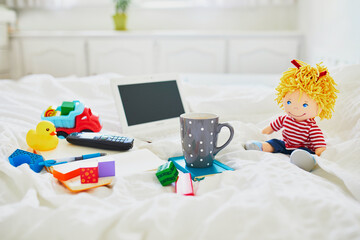 Laptop, cup of coffee, notebook, phone and different toys in bed on clean white linens