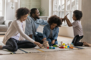 Fototapeta premium Happy African American family playing with toys, sitting on warm floor with underfloor heating, smiling parents with adorable little daughter and son building tower with wooden blocks cubes