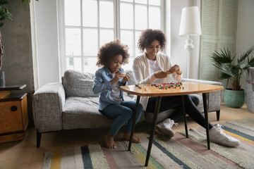 Smiling African American mother and daughter playing with colorful beads, sitting on couch at home,...