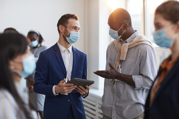 Waist up portrait of group of people chatting during break at business conference, focus on two men wearing masks with lens flare - Powered by Adobe