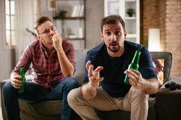 Two young friends enjoying at home. Men drinking beer and watching sports game on tv..