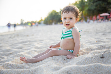cute toddler in a swimsuit looks at camera and sits on a sandy tropical beach of the warm sea in the sunshine