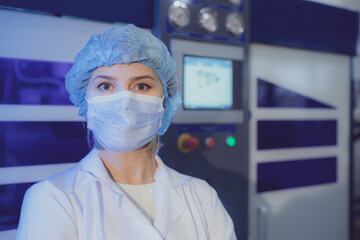 Woman employee with large eyes in medical mask hat and white uniform near equipment control panel in plant workshop closeup