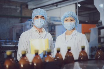 Young man and woman of middle age in a white coat, special caps and medical masks near a conveyor belt with filled bottles at a brewery