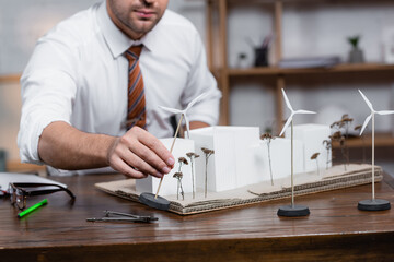 cropped view of architect holding model of wind turbine near architectural maquette, blurred background