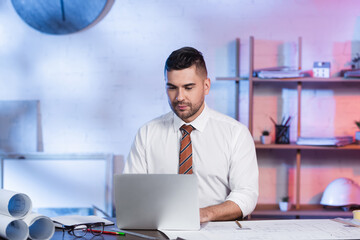 concentrated architect working on laptop near blueprints on desk