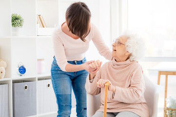 Kind woman helping lovely old lady to stand from chair