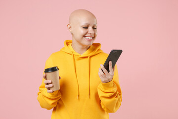 Young bald smiling caucasian friendly happy woman 20s without hair in yellow casual sweatshirt shirt hold plastic cup of coffee, mobile cell phone isolated on pastel pink background studio portrait.