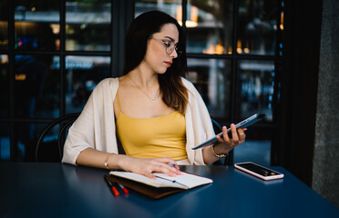 Caucasian blogger in glasses installing gadget application for online messaging and chatting, millennial hipster girl 20 years old booking tickets via touch pad during web networking in street cafe