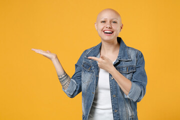 Young bald smiling happy caucasian woman 20s without hair in casual denim jacket white t-shirt point index finger aside on hand, workspace area mock up isolated on yellow background studio portrait.