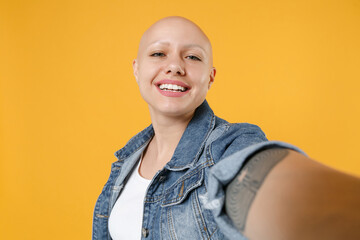 Close up young bald smiling happy friendly caucasian woman 20s without hair in casual denim jacket white t-shirt doing selfie shot on mobile phone isolated on yellow color background studio portrait.