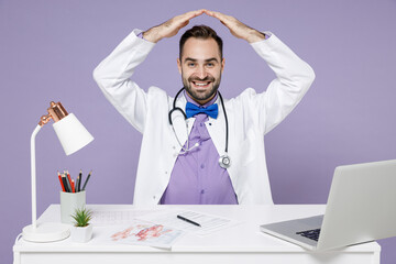 Male doctor man wearing white medical gown suit sit at desk work on computer in clinic office holding folded hands above head like roof, stay home isolated on violet color background studio portrait.