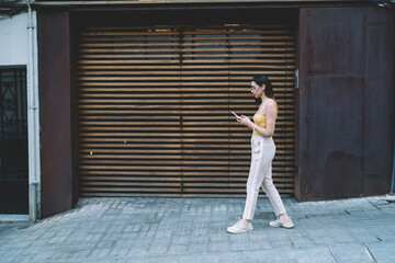 Fototapeta na wymiar Millennial blogger typing text for sharing content publication in social networks using smartphone device, side view of Caucaisan hipster girl writing email message while walking at urban street