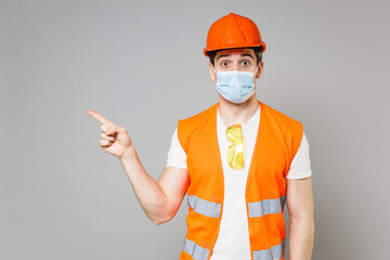 Young employee man in orange vest protective helmet face mask coronavirus point finger aside on workspace isolated on grey background studio Instruments for renovation apartment Repair home concept