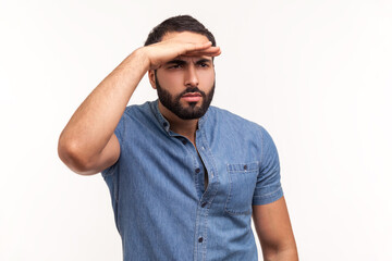 Thoughtful bearded man looking far away at distance with hand over head, attentively searching for...