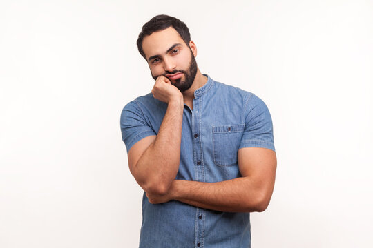 Bored upset man with beard in blue shirt leaning head on hand looking at camera with indifferent look, laziness and apathy, procrastination. Indoor studio shot isolated on white background