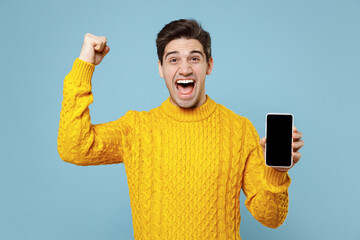Young happy excited man 20s wear knited yellow sweater hold in hand using mobile cell phone with blank screen workspace area do winner gesture clench fist isolated on blue background studio portrait.