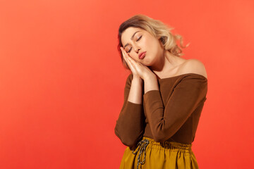 Fototapeta premium I want to sleep. Portrait of woman with blonde curly hair with closed eyes standing with arms under her head like pillow and sleep. Indoor studio shot isolated on red background