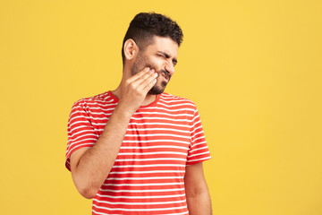 Unhappy bearded man in red striped t-shirt feeling toothache, touching sore cheek, suffering from...