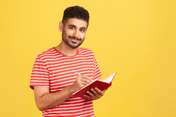Happy positive man with beard in striped t-shirt making notes with pen at notepad, writing down information and looking at camera with smile. Indoor studio shot isolated on yellow background