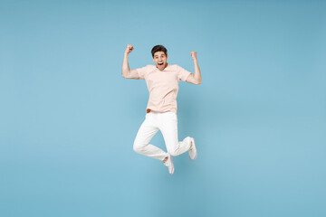 Fototapeta na wymiar Full length of young caucasian happy excited overjoyed student man 20s in beige t-shirt white pants doing winner gesture clench fists in air jumping high isolated on blue background studio portrait.