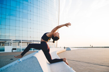 Experienced sportswoman with perfect body shape practicing pilates hatha yoga during leisure in city, inspired female enjoying morning workout for training stretching standing in Crescent pose