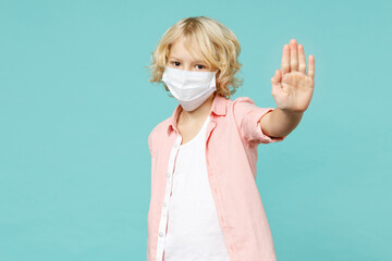 Little kid boy 10s in shirt sterile face mask safe from coronavirus virus covid-19 showing stop gesture with palm isolated on blue turquoise background studio portrait. Childhood lifestyle concept.