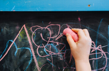 Little girl child draws with her right hand with pink chalk on a black wooden board, easel a...