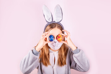 Girl in rabbit bunny ears on head and protective mask with colored eggs on pink background. Covid Easter holiday