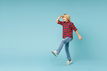 Full length side view excited little kid boy 10s in red checkered shirt holding hand at forehead looking far away distance isolated on blue background children portrait. Childhood lifestyle concept.