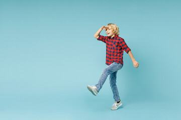 Obraz premium Full length side view excited little kid boy 10s in red checkered shirt holding hand at forehead looking far away distance isolated on blue background children portrait. Childhood lifestyle concept.